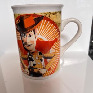 K&ouml;nnte beinhalten: Eine wei&szlig;e Keramiktasse mit einem farbenfrohen Bild der Figur Woody aus dem Animationsfilm Toy Story. Die Tasse hat einen gebogenen Henkel und ein Design, das sich um die Tasse wickelt.
