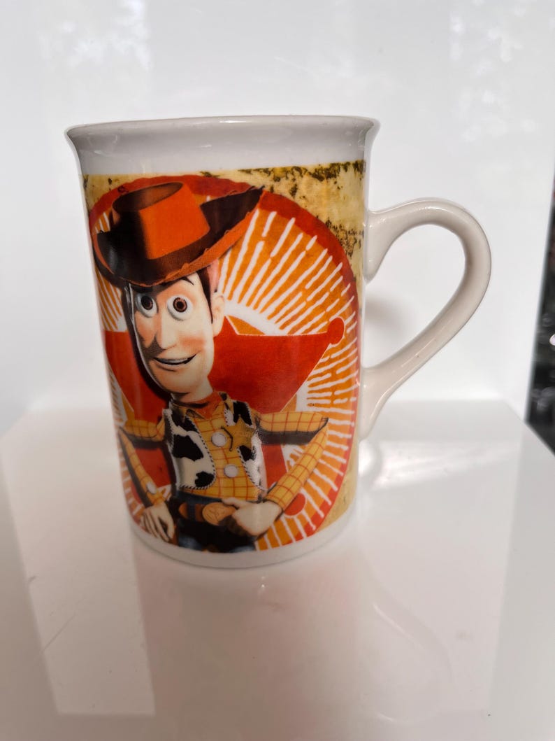 K&ouml;nnte beinhalten: Eine wei&szlig;e Keramiktasse mit einem farbenfrohen Bild der Figur Woody aus dem Animationsfilm Toy Story. Die Tasse hat einen gebogenen Henkel und ein Design, das sich um die Tasse wickelt.