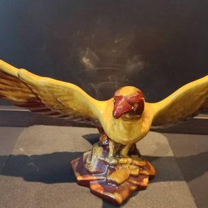 May include: A ceramic eagle figurine with outstretched wings, painted in shades of yellow and brown. The eagle is perched on a base with a textured, multi-coloured design.