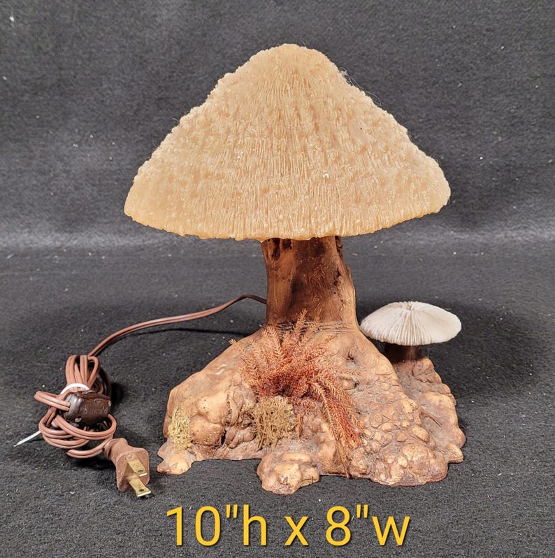May include: A mushroom-shaped lamp with a textured, beige lampshade and a brown base. The lamp is 10 inches high and 8 inches wide. A smaller mushroom is on the base, with brown and green accents. The lamp has a brown cord and plug.