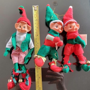 Peut inclure: Trois figurines de lutins de Noël. Les lutins portent des tenues rouges et vertes, avec des chapeaux et des chaussures rouges. Un lutin porte une chemise blanche et un gilet vert. Un mètre ruban est à côté des lutins, indiquant les mesures en centimètres.