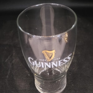 May include: A clear, tulip-shaped Guinness pint glass. The glass features the Guinness logo in gold and white, with a harp symbol and the text "ESTD 1759". The glass is empty.