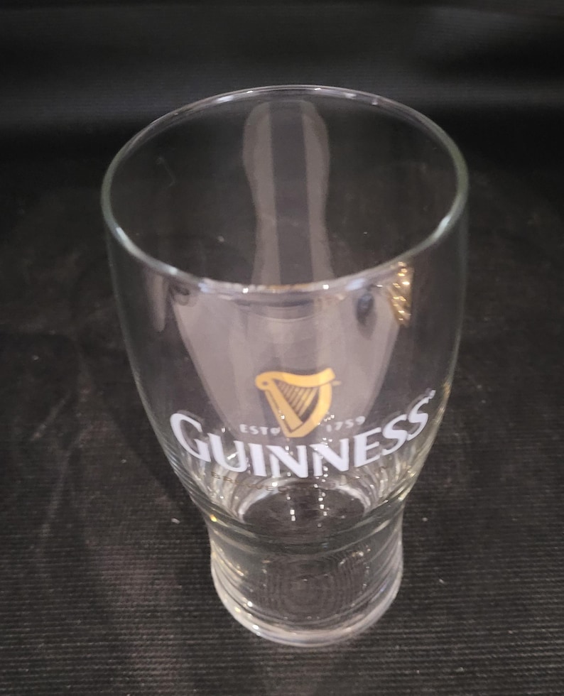 May include: A clear, tulip-shaped Guinness pint glass. The glass features the Guinness logo in gold and white, with a harp symbol and the text "ESTD 1759". The glass is empty.