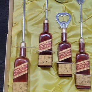 May include: A set of four miniature Johnnie Walker Red Label Scotch Whisky bottles, each with a different bar tool: a cocktail shaker, a bottle opener, a spoon, and a fork. The bottles are in a yellow satin-lined box.