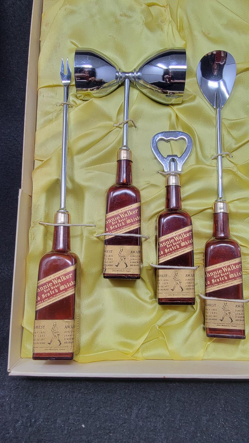 May include: A set of four miniature Johnnie Walker Red Label Scotch Whisky bottles, each with a different bar tool: a cocktail shaker, a bottle opener, a spoon, and a fork. The bottles are in a yellow satin-lined box.
