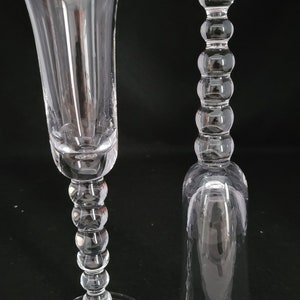 May include: Two clear glass champagne flutes with a beaded stem design. The flutes are on a black background.