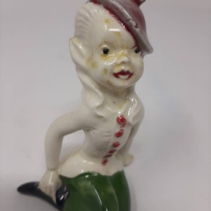 May include: A ceramic figurine of a kneeling elf wearing a white shirt with red buttons, green trousers, and a red and white hat.