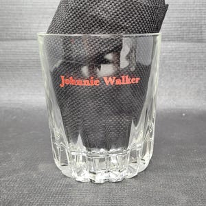 May include: A clear, faceted glass tumbler with the red text "Johnnie Walker" on the front. The glass is partially filled with a black, textured material. The glass has a thick base and a slightly flared rim.