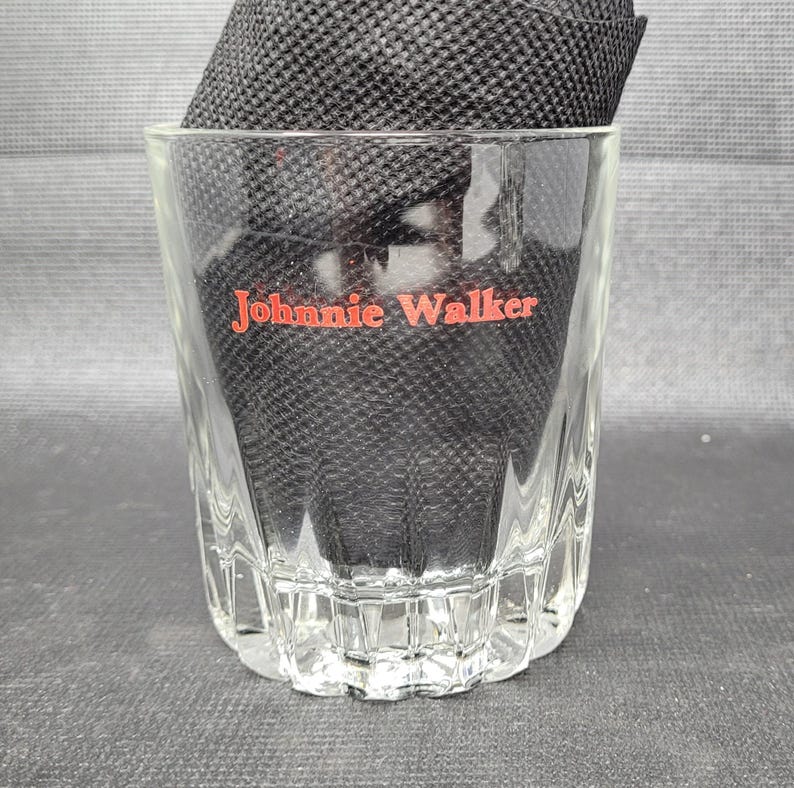 May include: A clear, faceted glass tumbler with the red text "Johnnie Walker" on the front. The glass is partially filled with a black, textured material. The glass has a thick base and a slightly flared rim.