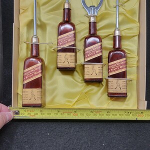 May include: A set of four miniature Johnnie Walker Scotch Whisky bottles with a cocktail shaker, a bottle opener, a spoon, and a cocktail fork. The bottles are in a yellow box with a gold lining.