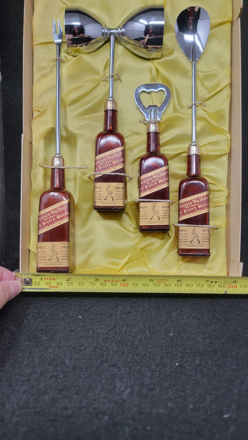 May include: A set of four miniature Johnnie Walker Scotch Whisky bottles with a cocktail shaker, a bottle opener, a spoon, and a cocktail fork. The bottles are in a yellow box with a gold lining.