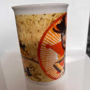 K&ouml;nnte beinhalten: Wei&szlig;er Keramikbecher mit einem Cartoon-Cowboy-Design. Der Becher hat einen beigefarbenen, strukturierten Hintergrund und ein orange-wei&szlig;es kreisf&ouml;rmiges Design. Der Cowboy tr&auml;gt einen Hut und eine Weste. Geeignet f&uuml;r Hei&szlig;getr&auml;nke.