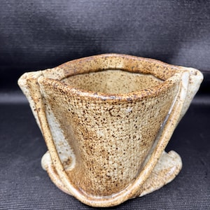 Peut inclure: Un verseur à café en céramique fait à la main avec un glaçage beige moucheté. Le verseur a un design angulaire unique avec une large ouverture et une base incurvée. Le bord et les bords sont brun foncé, ajoutant du contraste.