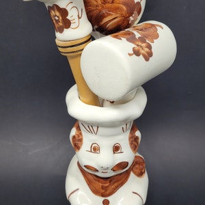 Może przedstawiać: Ceramiczna figurka kucharza z białą podstawą i brązowymi detalami malowanymi. Kucharz trzyma zestaw przyborów kuchennych, w tym solniczkę w kształcie grzyba, okrągłą pieprzniczkę i łyżkę z drewnianym uchwytem.