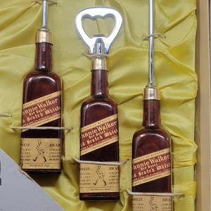 May include: A set of three miniature bottles of Johnnie Walker Red Label Scotch Whisky, each with a bottle opener and a silver cocktail spoon. The bottles are displayed on a yellow satin background.