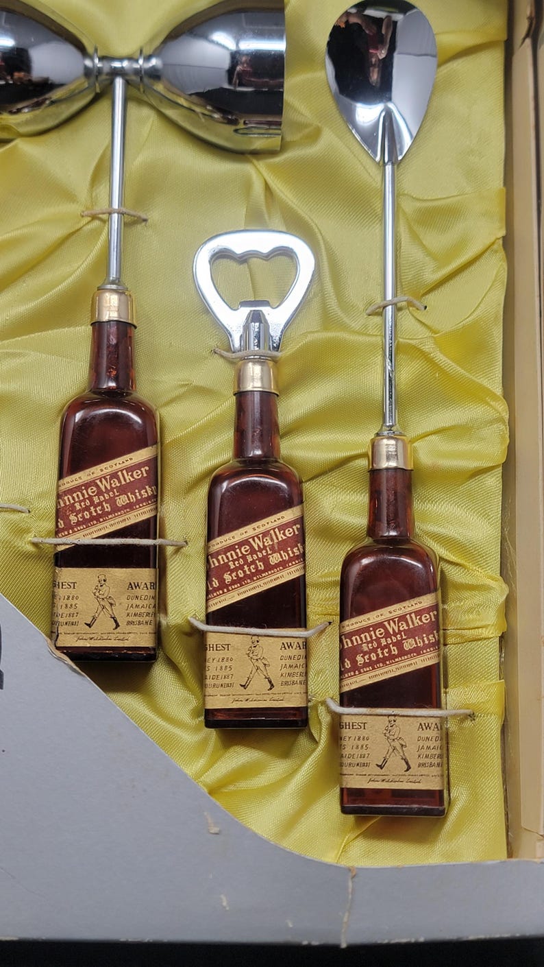 May include: A set of three miniature bottles of Johnnie Walker Red Label Scotch Whisky, each with a bottle opener and a silver cocktail spoon. The bottles are displayed on a yellow satin background.