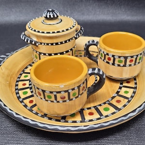 May include: A decorative ceramic tea set with a tray. The set includes two small mugs, a lidded sugar bowl, and a round tray. The pieces are beige with black, red, green, and yellow accents. The tray has a black and white patterned border.