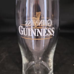May include: A clear, tulip-shaped Guinness pint glass. The glass features the Guinness logo in white, with a gold harp emblem above the brand name. The glass is empty and appears to be clean.