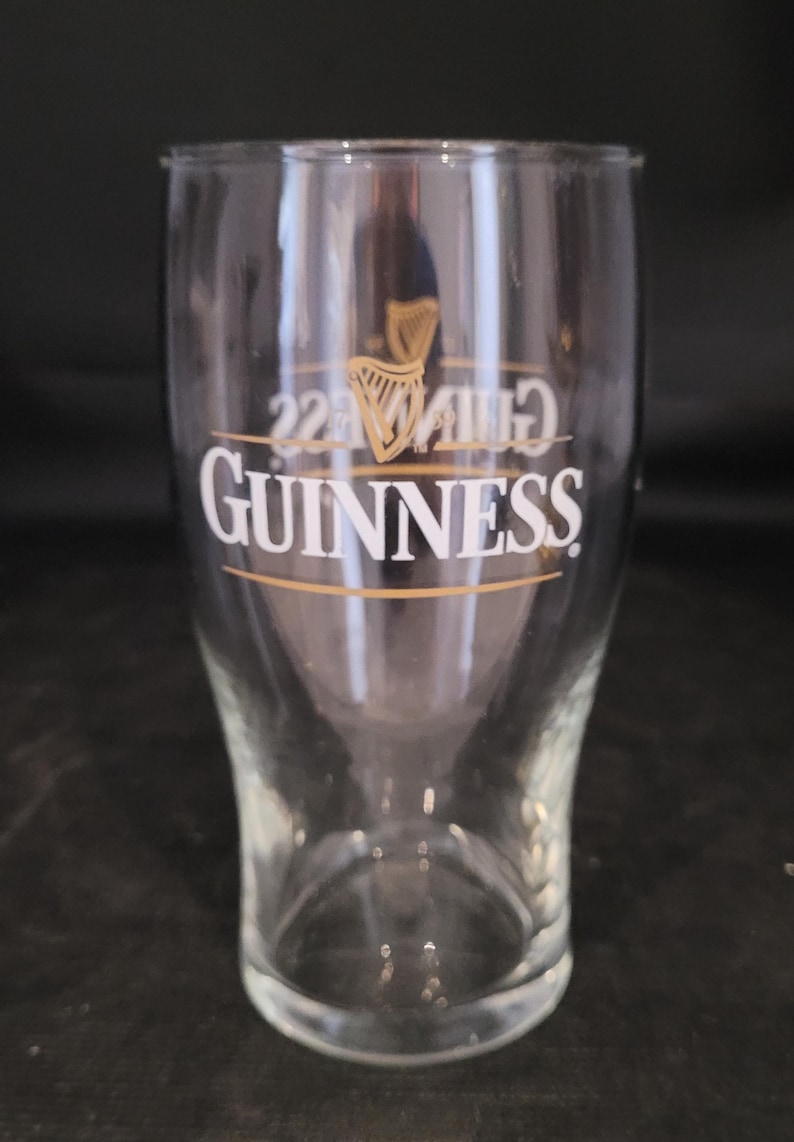 May include: A clear, tulip-shaped Guinness pint glass. The glass features the Guinness logo in white, with a gold harp emblem above the brand name. The glass is empty and appears to be clean.