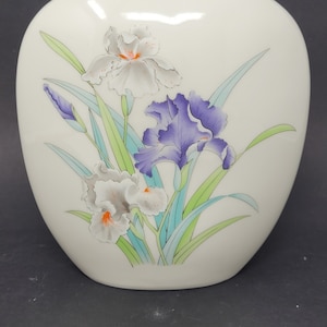 May include: White ceramic vase with a floral design featuring three irises. The irises are painted in shades of purple, white, and orange. The vase has a rounded shape and a smooth, glossy finish.