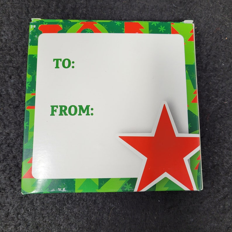 May include: A square gift tag with a white centre and a red star. The tag has the words "TO:" and "FROM:" in green. The border is green with a repeating Christmas tree pattern.