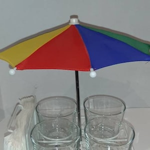 May include: A white metal drink holder with four clear glass cups and a colourful umbrella. The holder has a wheel on the bottom and a napkin holder on the side.