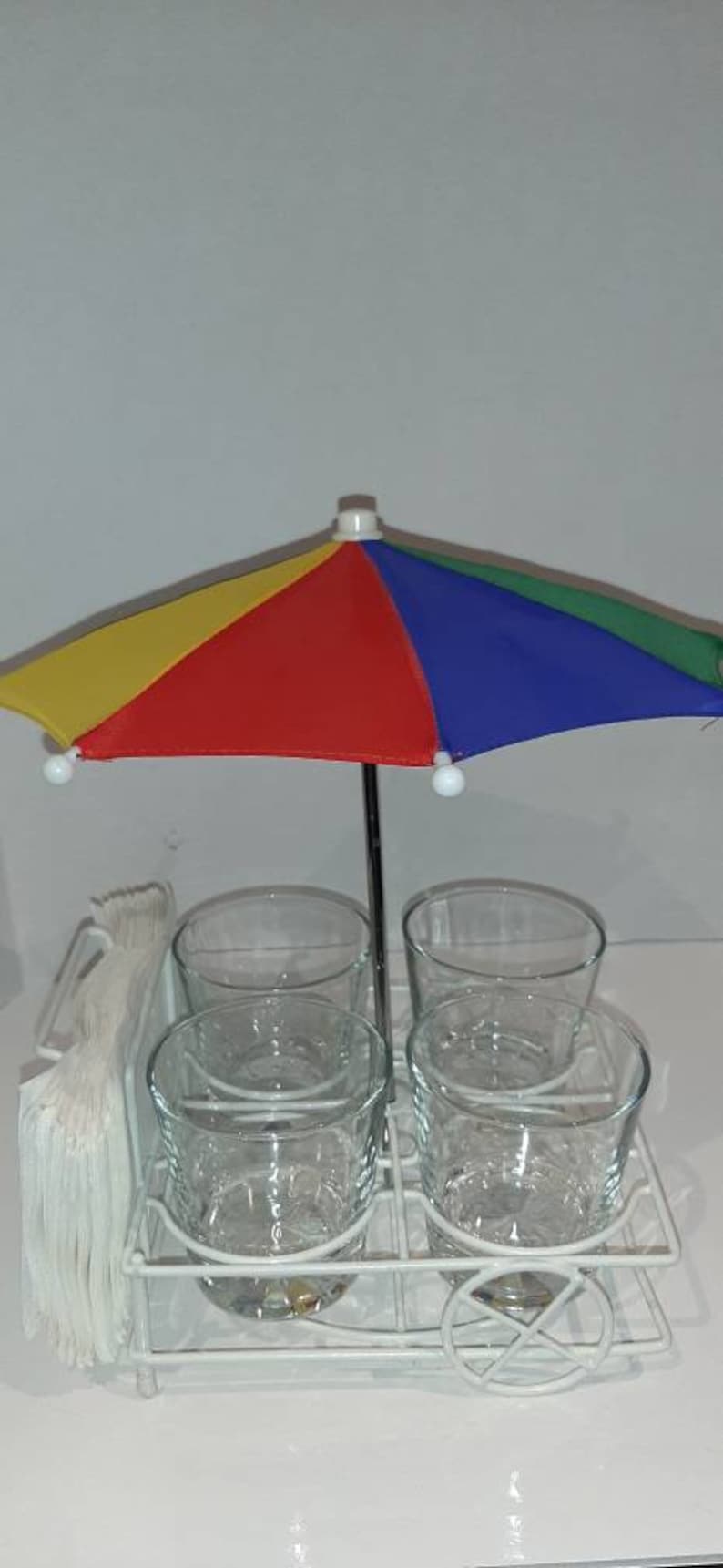 May include: A white metal drink holder with four clear glass cups and a colourful umbrella. The holder has a wheel on the bottom and a napkin holder on the side.