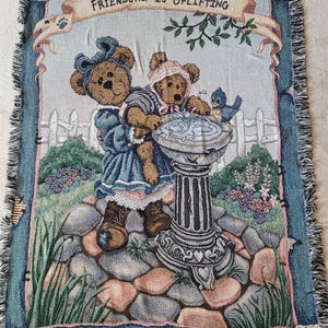 May include: A woven tapestry blanket, 150cm high x 132cm wide, depicts two teddy bears near a fountain, with a blue bird. The text "FRIENDSHIP IS UPLIFTING" is displayed on a banner. The blanket has a fringed edge and the text "BOYDS BEARS & FRIENDS".
