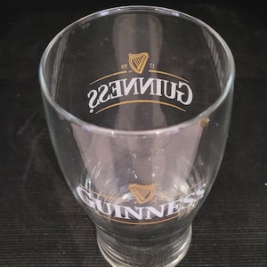 May include: A clear, tulip-shaped Guinness pint glass. The glass features the Guinness logo in gold and white, with the word "Guinness" and a harp emblem prominently displayed. The glass is empty and sits on a dark surface.