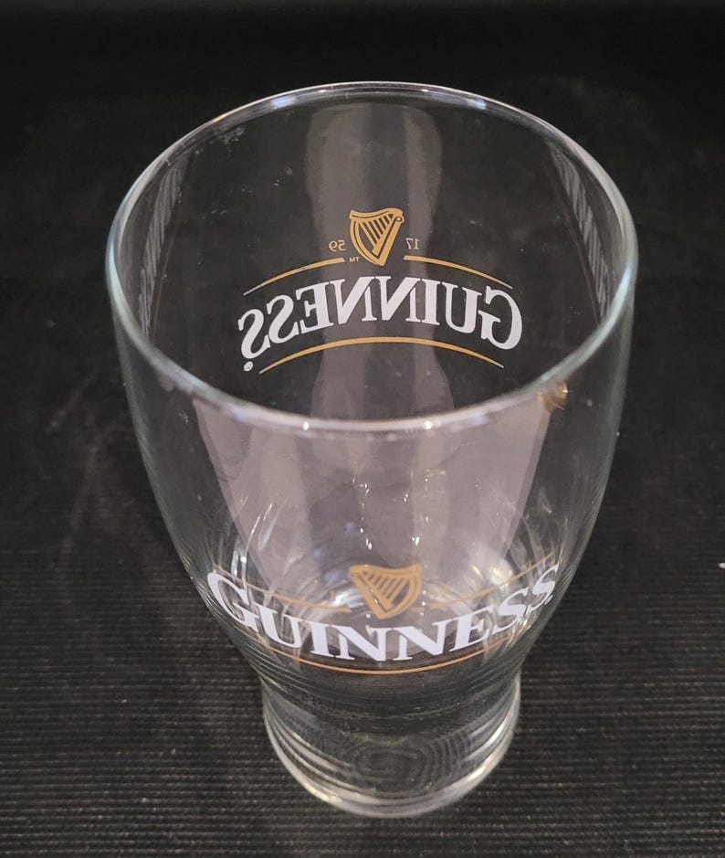 May include: A clear, tulip-shaped Guinness pint glass. The glass features the Guinness logo in gold and white, with the word "Guinness" and a harp emblem prominently displayed. The glass is empty and sits on a dark surface.