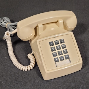 Peut inclure: Un téléphone filaire beige avec un clavier. Le téléphone comprend un combiné, une base avec un clavier et un cordon spiralé. Le clavier comporte des boutons noirs avec des chiffres et des lettres blancs. Le téléphone est sur une surface sombre.