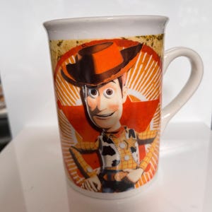 K&ouml;nnte beinhalten: Wei&szlig;er Keramikbecher mit einem Cartoon-Bild von Woody aus Toy Story. Das Design zeigt Woodys Gesicht, Hut und Outfit vor einem roten und orangefarbenen Sternenhintergrund. Der Becher hat einen gebogenen Griff.