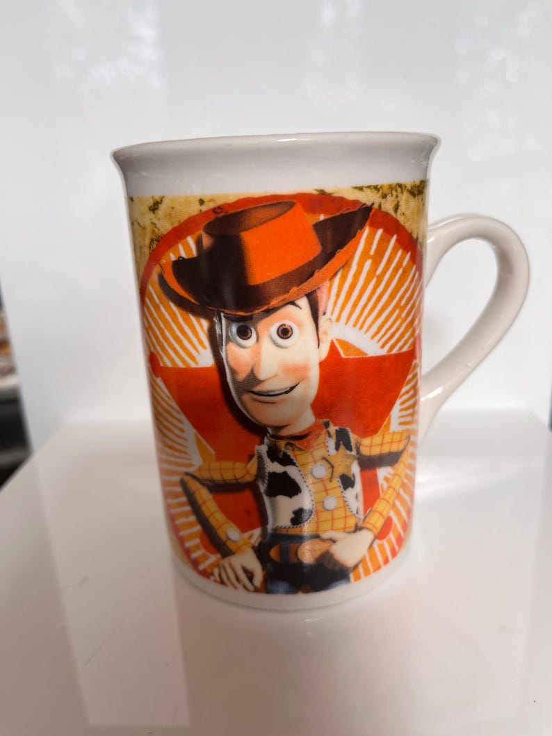 K&ouml;nnte beinhalten: Wei&szlig;er Keramikbecher mit einem Cartoon-Bild von Woody aus Toy Story. Das Design zeigt Woodys Gesicht, Hut und Outfit vor einem roten und orangefarbenen Sternenhintergrund. Der Becher hat einen gebogenen Griff.