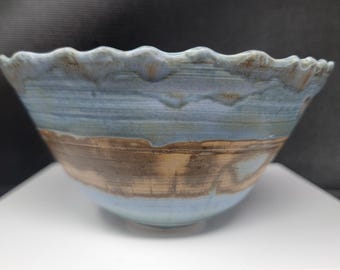 May include: A handmade ceramic bowl with a wavy rim. The bowl features a light blue glaze on the upper portion and a brown band around the middle. The interior is a light blue color. The bowl is sitting on a white surface.