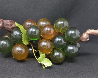May include: A glass grape cluster with green and amber grapes on a brown wooden vine. The grapes are arranged in a bunch and are realistic looking.