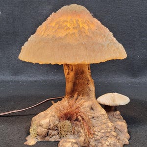 May include: A mushroom-shaped lamp with a textured, tan-colored cap that glows with warm light. The lamp's stem and base are designed to resemble a tree trunk, with detailed textures and small decorative mushrooms. A brown power cord extends from the base.
