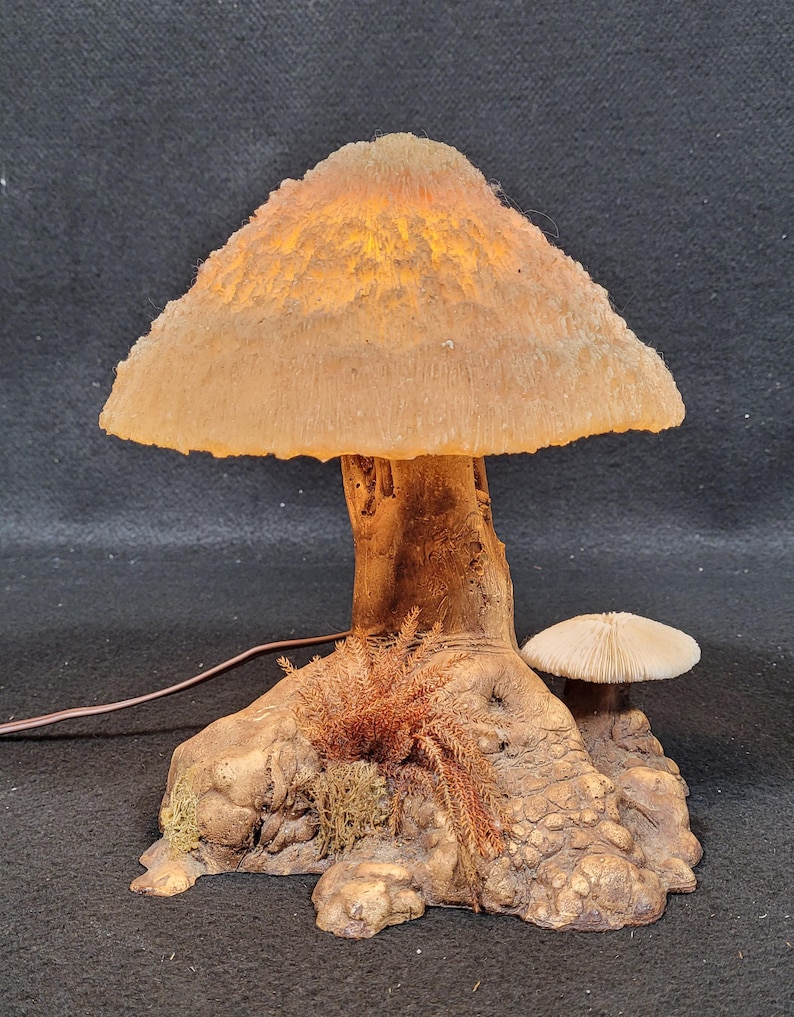 May include: A mushroom-shaped lamp with a textured, tan-colored cap that glows with warm light. The lamp's stem and base are designed to resemble a tree trunk, with detailed textures and small decorative mushrooms. A brown power cord extends from the base.