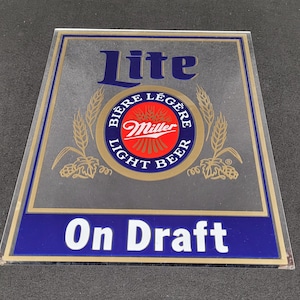 May include: A rectangular sign with a clear background and a blue border. The sign features the word "Lite" in blue, a circular logo with "Miller" in red, and the words "On Draft" in white on a blue banner.