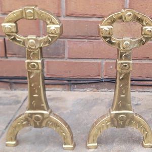 Peut inclure: Deux chenets de cheminée antiques en laiton doré. Chaque chenet présente un dessus ovale décoratif, une section centrale rectangulaire et une base incurvée avec un motif floral. Ils sont posés sur une surface grise devant un mur de briques.