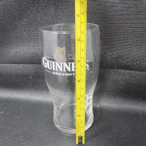 May include: A clear Guinness pint glass with the Guinness logo and the word "DRAUGHT" printed on the front. The glass is approximately 7 inches tall, as measured by a yellow tape measure.