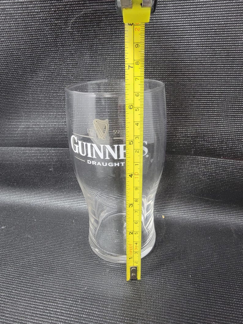 May include: A clear Guinness pint glass with the Guinness logo and the word "DRAUGHT" printed on the front. The glass is approximately 7 inches tall, as measured by a yellow tape measure.