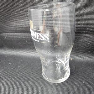 May include: A clear, empty Guinness pint glass with the Guinness logo in white. The glass has a curved shape and a wide base. The glass is on a black surface.