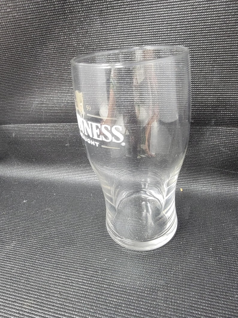 May include: A clear, empty Guinness pint glass with the Guinness logo in white. The glass has a curved shape and a wide base. The glass is on a black surface.
