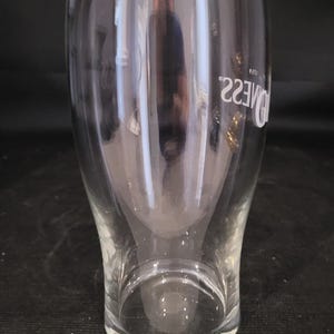 May include: A clear, tulip-shaped pint glass with the word "Guinness" printed in white. The glass has a wide top, a curved body, and a sturdy base. The glass is designed for serving beer.