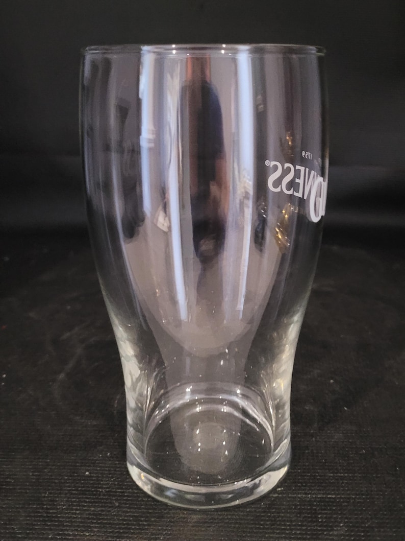 May include: A clear, tulip-shaped pint glass with the word "Guinness" printed in white. The glass has a wide top, a curved body, and a sturdy base. The glass is designed for serving beer.