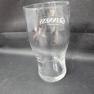 May include: A clear, tulip-shaped pint glass with the Guinness logo in white. The logo includes the word "Guinness" in a stylized font and a harp symbol. The glass is empty and sits on a dark surface.