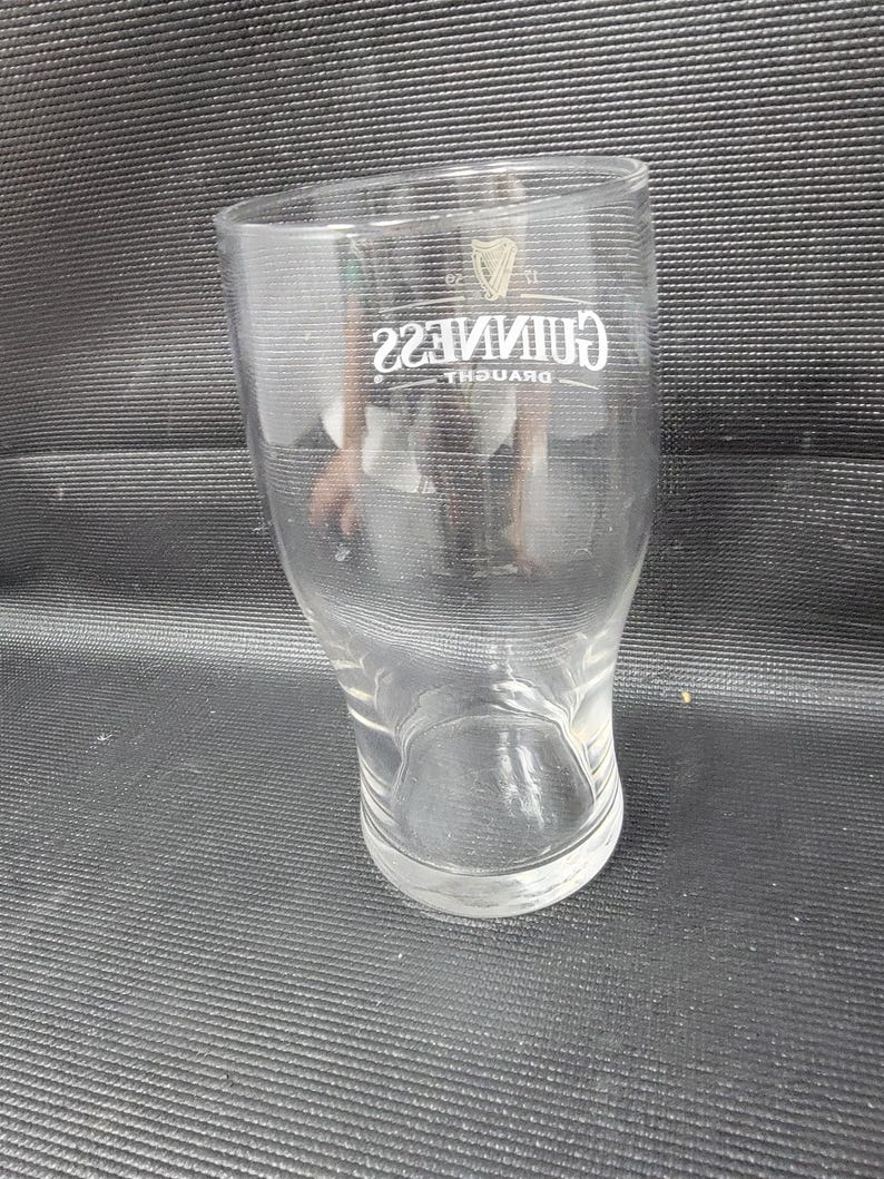 May include: A clear, tulip-shaped pint glass with the Guinness logo in white. The logo includes the word "Guinness" in a stylized font and a harp symbol. The glass is empty and sits on a dark surface.