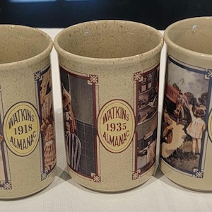 May include: A set of six beige ceramic mugs. Each mug features a framed image and a circular emblem with a year, including 1914, 1916, 1918, 1933, 1936, and 1939. The images depict historical scenes.