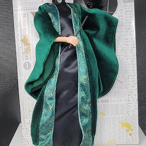 May include: A doll of a character from the Wizarding World, wearing a green velvet robe with patterned trim over a black dress. The doll has blonde hair and is holding a wand. The packaging includes the text "Wizarding World".