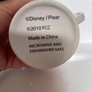 K&ouml;nnte beinhalten: Wei&szlig;er Keramikbecher mit Henkel. Der Boden des Bechers zeigt den Text "&copy;Disney / Pixar", "&copy;2010 FCC", "Made in China" und "MICROWAVE AND DISHWASHER SAFE" in schwarzer Schrift.